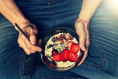 man sitting cross-legged eating a bowl of healthy grains topped with fresh fruit and nuts - foods