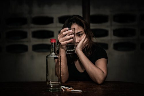 young woman looking at a glass of colorless liquor while sitting a table with an empty bottle and several cigarettes - meth and alcohol